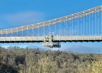 A small, movable cage with two persons standing in it, is hanging below a large white suspension bridge