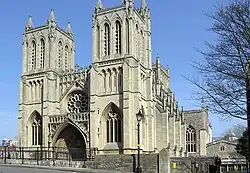 A stone-built Victorian Gothic building with two square towers and a central arched entrance underneath a circular ornate window. A Victorian street lamp stands in front of the building and on the right part of a leafless tree, with blue skies behind.