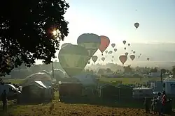 A large number of hot air balloons taking off from a field which is surrounded by tents and stalls. The sun is low in the sky, and balloons can be seen flying into the distance.