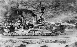 Overhead view of a large battleship; black smoke pours from its smoke stacks as it steams through choppy seas.