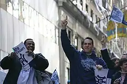 A black man on the left wearing a white T-shirt and dark jacket smiles holding a sign, while a white man on the right wearing a blue sweatshirt which reads "Champions" points upwards. White streamers hang from a building in the background.