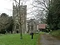 Square grey tower of stone church building, partially obscured by trees. Red roofed lych gate to right. Grass and gravestones in the foreground