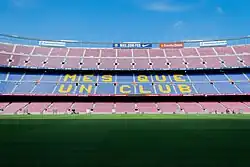 The words "Més que un club" are painted in yellow on the blue seats of the stadium