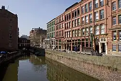 A canal as photographed from a bridge over it. On the left hand side there is a brick industrial building that is in shadow; on the right hand side there are brick buildings with many windows facing over the canal, which us lined by brick wall.