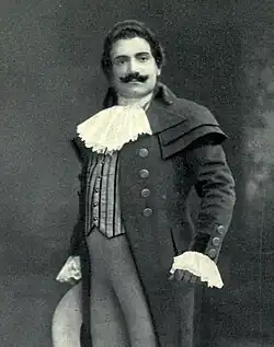 Man, with dark hair and a curling moustache, standing in a posed position. He is wearing a long coat, with lace at the throat and cuffs.