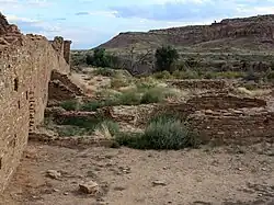 A partly overcast sky and subdued sunlight over a roughly six-foot tall wall of dusky tan sandstone bricks that vary somewhat in size. The wall runs diagonally from the immediate foreground at left toward the right, running perhaps several dozen feet to the near middle distance. A few feet to the right, in the middle foreground, a low ring of similar blocks delimits a circular pit sunk into the ground. The remains of several other ruinous low walls, perhaps one to three high at most, are arrayed in parallel; they align left to right from the high diagonal wall. Perhaps a mile distant to the center and right, a canyon wall slopes gradually level to meet the valley floor on which the walls sit.