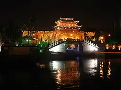 A traditional Chinese city gate and its surrounding wall viewed at night, brightly lit from below in warm, golden light. A curved stone bridge spans the dark water in the foreground, with the gate tower standing prominently atop the wall.