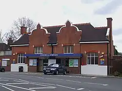 A brown-bricked building with a black roof and a blue sign that reads "CHIGWELL STATION" in white letters all under a white sky