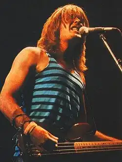 Williams, about 31, shown in upper body shot. He sings into microphone on a stand while strumming his guitar. He wears a singlet top and has shoulder length hair.