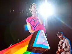 A man wearing a pink shirt sings with a microphone while holding a flag
