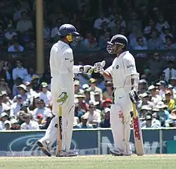Two men wearing white uniforms shirts and trousers, blue helmets, gloves, pads and holding cricket bats meet one another on a cricket pitch to touch gloves. A crowd can be seen in the distance.
