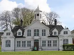 Ornate, symmetrical, wooden house with two smaller wings with decorative engaged columns. The pitched roof has gables. The windows are paned and the front door has columns on either side.