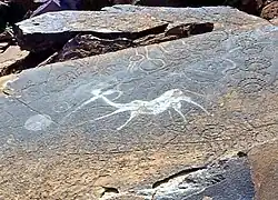 A sandstone slab in horizontal position covered by a dark patina that appears greasy black as if dirtied by touch. In the middle of the slab is a clear white indentation of a fantasy creature with the body of an antelope and the torso and head of a human who appears to b shooting at something with a bow. The figure is surrounded by other engravings that appear geometric and have the color of the slab surface.