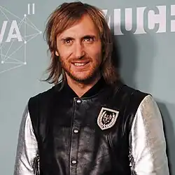 Image of a man with long hair and a beard, wearing a black and silver jacket, standing in front of a patterned backdrop.