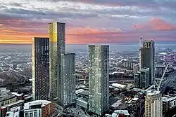 An aerial photo of five glass and steel skyscrapers at the centre of a city with a large urban sprawl stretching the entire background. The sky is cloudy with the sunset creating an orange hue reflected by the buildings' glass exteriors.