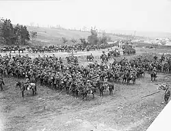 A large group of men and horses drawn up into lines in a field and on the adjoining road. A hill with trees and tents can be seen in the background.