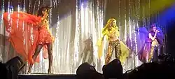 Members of the group each dressed in colorful costumes—red, yellow, and purple. They are performing a dance routine under stage lighting, with a shiny backdrop behind them.