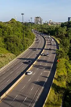 A photo of a freeway in a ravine in the autumn, taken from atop the edge of the ravine. Projecting above the forest is the top of several skyscrapers and the CN Tower.