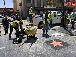 Construction workers in high-vis yellow vests work with trash bags and a wheelbarrow on the sidewalk around a recently poured terrazzo pad and unidentifiable star. Police officers in front of caution tape and pedestrians behind caution tape watch. Disney, Ghirardello, and TMZ banners are visible across the street.