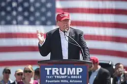 Trump speaking in front of an American flag behind a lectern, wearing a black suit and red hat. The lectern sports a blue "TRUMP" sign.