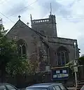 Red and grey stone building with arched windows and triangular roof. Behind is a small square tower