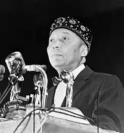 A clean-shaven, middle-aged black man, wearing an embroidered hat, standing at a podium with microphones in front of him.
