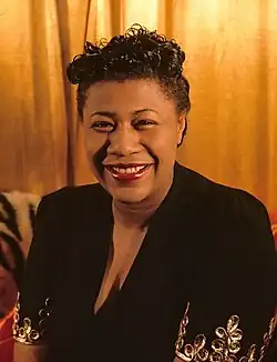 Headshot of Ella Fitzgerald. She was a black woman with dark curly hair. She is a wearing a black dress while standing in front of a golden curtain smiling