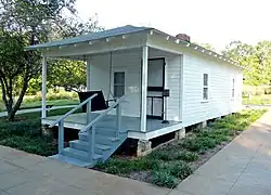 Present-day photograph of a whitewashed house, about 15&nbsp;feet wide. Four banistered steps in the foreground lead up to a roofed porch that holds a swing wide enough for two. The front of the house has a door and a single-paned window. The visible side of the house, about 30&nbsp;feet long, has double-paned windows.