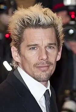 Hawke with bleached hair, wearing a black overcoat. The collar of a gray suit is visible in the photo, with a white shirt and dark gray tie.