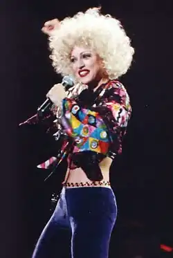 Madonna with short blonde hair, wearing a green bra and purple pants, singing to a microphone, held in her left hand.