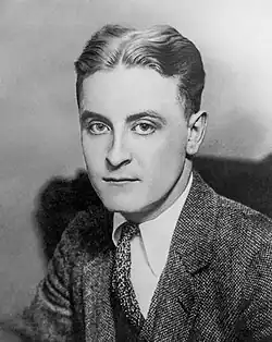 A photograph of F. Scott Fitzgerald by Nickolas Muray. Fitzgerald is bent over a desk and is examining a sheaf of papers. He is wearing a light suit and a polka-dot tie. A white handkerchief is in his breast pocket.