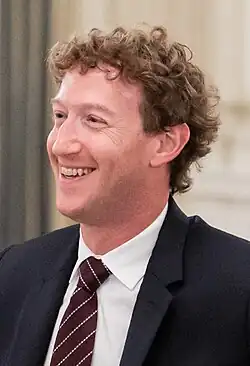 Side view of Zuckerberg smiling and wearing a suit with a striped tie