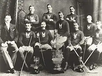 Two rows of ice hockey players, with the front row seated and most of the back row standing. Some of the players are holding hockey sticks, and various trophies are placed in front of them.