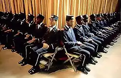 A group of black men, wearing navy blue uniforms, seated in two lines which intersect to form a V formation.