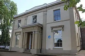 A small stucco building with sash windows. It is a cube shape with a slightly slanted roof and two stories, as photographed from the ground in front. Trees are seen on either side of the building.