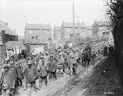 a large crowd of German soldiers walking along a small road under the supervision of soldiers wearing British equipment on horseback. Houses can be seen in the background.
