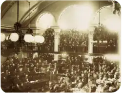 A photograph showing a large number of men seated on semi-circular tiers in a vaulted chamber as a large crowd looks on from an arcaded balcony