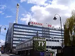 A glass and steel building that is a cuboid shape with a perpendicular single-storey extension sticking out towards the camera. The side of this extension closest to the camera is decorated with leaves and has a white sign that read "GRANADA / TV"