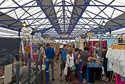 An interior of a building with a translucent glass roof supported by blue-painted steel latticework. On the main floor are a number of different stalls with customers inspecting various wares.