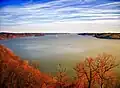 View of a very wide river lined with bare trees in reddish light under a blue sky streaked with clouds