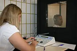 a female medical professional is seated in front of a special sound-proof booth with a glass window, controlling diagnostic test equipment. Inside the booth, a middle-aged man can be seen wearing headphones and is looking straight ahead of himself, not at the audiologist, and appears to be concentrating on hearing something
