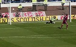 A football player scores a goal against the opposing goalkeeper from a penalty-kick. Stewards and camera-operators are visible behind the goal net.
