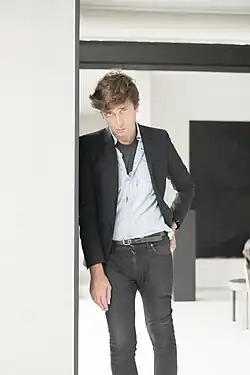 A man with tousled hair leans casually against a wall, wearing a blazer over a light shirt and dark jeans in a modern, minimalist room