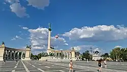 A wide-angle view of Heroes Square in Budapest showing the Millennium Monument, colonnades, and visitors in the open plaza under a clear sky.