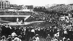 Wide shot of a black-and-white photograph of a baseball field, with spectators in the foreground and background.
