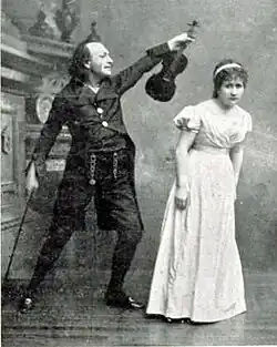 Photograph of maniac in black 19th century day clothes brandishing a violin at a frightened young woman in a full-length white frock