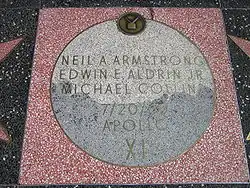 Moon Landing monument, with square pink terrazzo pad, with half light, half medium-dark gray terrazzo Moon disk showing TV emblem at top and brass lettering inlaid that reads: Neil A Armstrong / Edwin E Aldrin Jr / Michael Collins / 7/20/69 / Apollo / XI