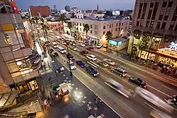 Downward view on a well-trafficked street surrounded by mid-rise buildings. On one side of the street are ceremonial stars embedded in a polished sidewalk.
