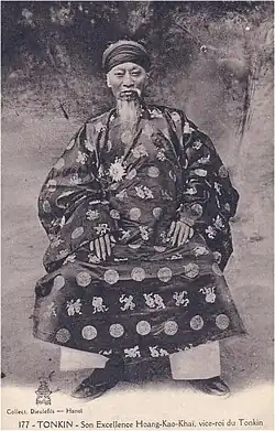 Aged man with cylindrical headdress, long flowing beard and moustache, a traditional Vietnamese tunic, darkly coloured with a light circular and Chinese character imprints, light coloured trousers, and dark shoes, sitting in a chair.