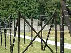 Taken from outside the reconstruction of a barracks, the photo shows a barbed wire fence, and beyond it a grassy area with a small timber hut